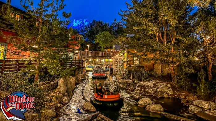 Rafts splash along a rocky waterway beneath trees and warm lights at dusk.